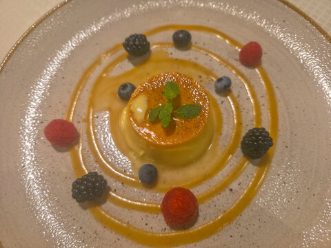 Creamy caramel flan plated with concentric caramel sauce rings, fresh raspberries, blackberries and blueberries, topped with mint leaves; an elegant, ideal for restaurant menus or food blogs.