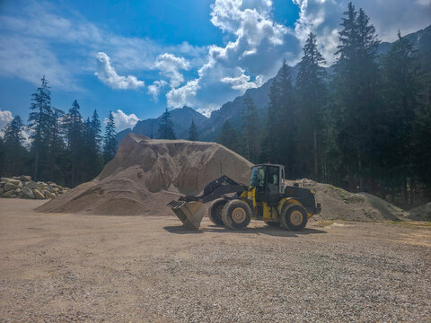 Yellow front loader parked beside a large gravel pile at a mountain quarry, framed by pine trees and dramatic clouds, conveying heavy industry blending with rugged natural landscape.