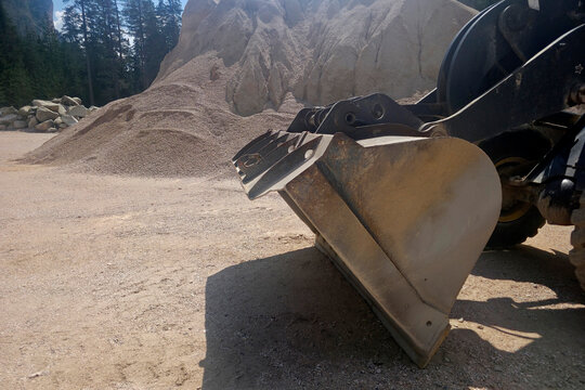 Yellow front loader parked beside a large gravel pile at a mountain quarry, framed by pine trees and dramatic clouds, conveying heavy industry blending with rugged natural landscape.