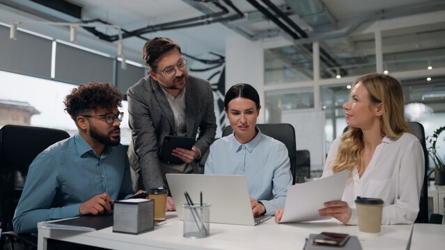 Four multiethnic business managers talking laughing teamwork using laptop reviewing papers documents stats result statistic legal data multiracial businesspeople diversity men women talk work briefing