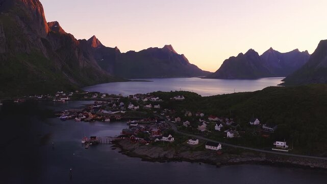 An aerial view captures the coastal village of Reine in Lofoten Norway surrounded by calm fjord waters and dramatic mountain peaks at dusk.