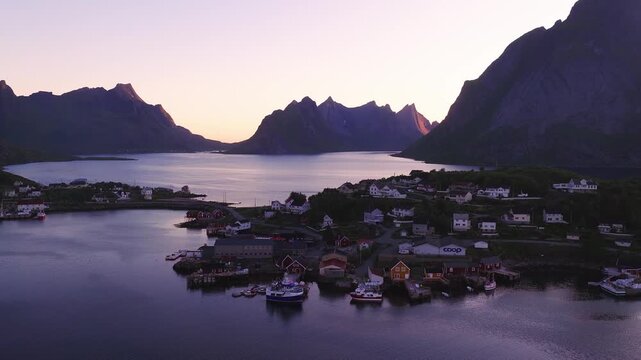 Aerial view over the coastal village of Reine in Lofoten Norway with traditional houses, boats at the pier, and mountains during sunset.