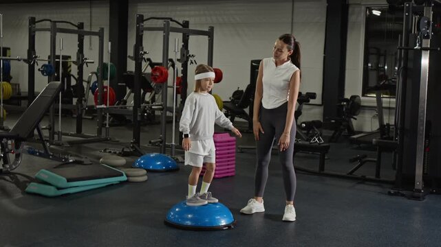 Caucasian female trainer supporting Caucasian male child balancing on one leg on balance trainer in gym. Clip suitable for marketing youth fitness, physical therapy, or family wellness campaigns