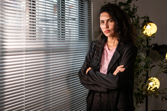 Confident businesswoman standing beside office blinds, looking at the camera