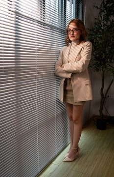 Confident businesswoman standing beside office blinds, looking at the camera while working