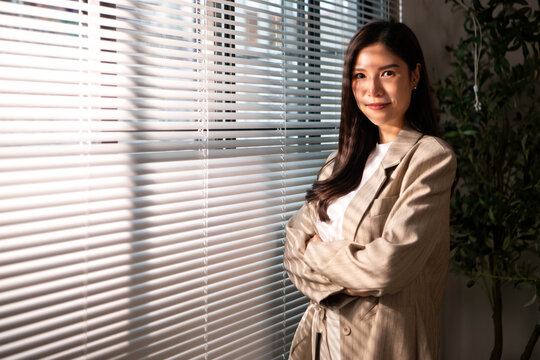 Confident businesswoman standing beside office blinds, looking at the camera while working