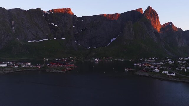 An aerial perspective shows Reine village in Lofoten, Norway, set against steep rocky mountain ridges illuminated by golden morning light beside the fjord waters.