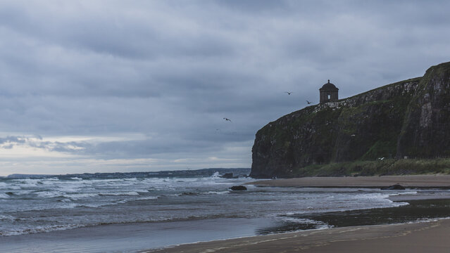 Coleraine, Northern Ireland, United Kingdom - 03 September 2023: View of Mussenden Temple perched on a cliff overlooking the sandy beach and crashing waves under a cloudy sky.