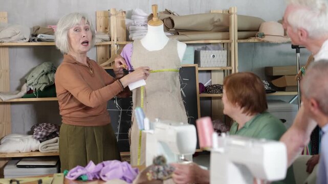  Sewing school senior woman teacher conducts lesson for elderly students, using mannequin to explain principle of constructing product, connecting parts of dress with back and front seams.