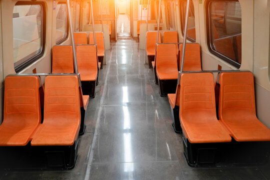 Bright afternoon sunlight shines into an empty subway train interior showing rows of orange plastic seats and a reflective floor inside a quiet city transport car