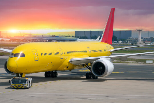 Service tug truck pushes a bright yellow cargo airplane on the tarmac at an international airport while the sun sets behind the terminal buildings
