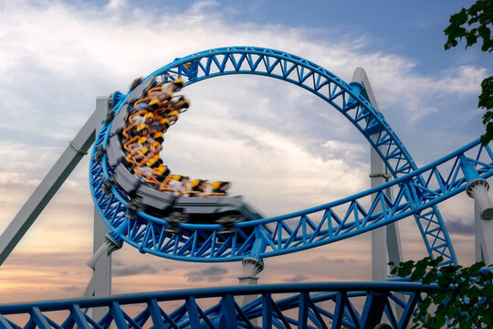 People ride a bright blue roller coaster while moving through a loop track at a park during the evening hours with a cloudy sky in the background