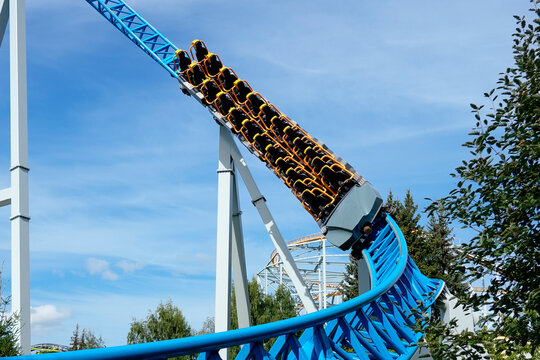 Empty seats test ride a high-speed blue roller coaster train along a steep metal track at an outdoor theme park on a sunny day.