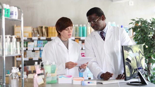 Two pharmacist employee using computer, reads service protocol update, finds out changes, and becomes familiar with work regulations. Woman and African man pharmacists at PC