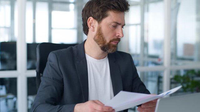 Caucasian middle-aged business man serious concentrated reading papers typing laptop 40s businessman read documents review data analysing work reports stats mature male working with computer in office