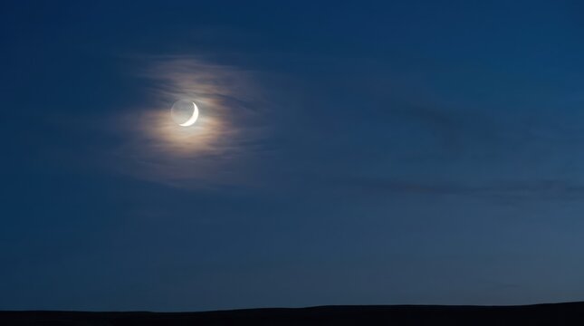 Crescent moon partially obscured by thin clouds during twilight hours