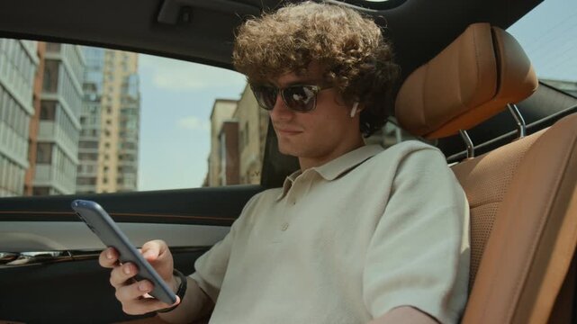 Caucasian young adult man sitting in backseat of moving car, using smartphone and wearing wireless earbuds. Video is suitable for ride-sharing apps, telecom ads, or mobile connectivity promos