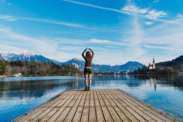 Fototapeta premium A young woman makes a heart gesture with her hands on a wooden pier at Lake Bled, Slovenia. Joyful travel lifestyle concept captures love, positivity, and freedom in scenic nature.