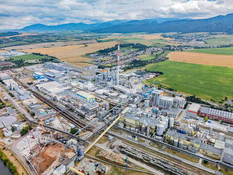 Aerial view of the Mondi SCP factory complex with its tall chimney, industrial buildings, and surrounding green fields and mountains in Ruzomberok, Zilinsky kraj, Slovakia.