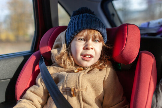 Cute young child with red hair wearing hat and coat, sitting securely in red car seat during daytime trip. Safety and travel, childhood, family vacation, transportation, protection, journey.