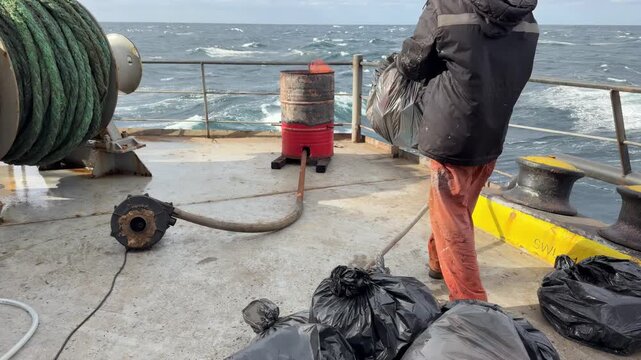Ship crew member incinerating sorted trash in a barrel on deck. Cargo vessel navigating through the open blue sea waves with visible fire and smoke.