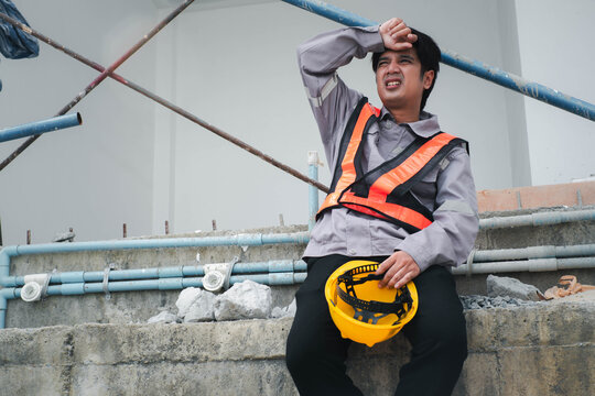 Tired construction worker in a reflective safety vest sits at a building site holding a hard hat, showing fatigue, physical labor, workplace safety, stress, exhaustion, and construction work.
