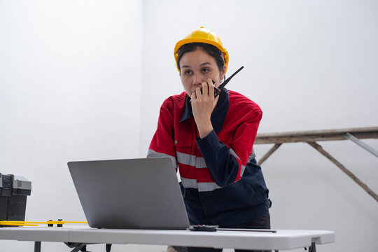 Female construction worker in a hard hat uses a two-way radio beside a laptop at a worksite, representing field communication, digital project management, engineering, and modern industrial operations