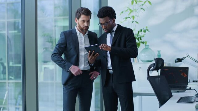 Two multiethnic business men talking looking in digital tablet device discussing work startup online project in office multiracial businessmen males agents talk working together with wireless gadget