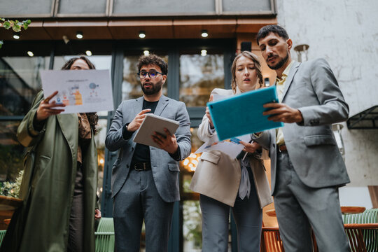Business colleagues review documents and tablets while standing outside an urban cafe. A small group discusses ideas during an outdoor meeting using paper charts and digital devices.