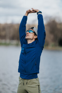 A young girl in a blue jacket and sunglasses stretches her arms overhead beside a calm lake. The outdoor fitness scene shows a girl warming up before exercise in cool weather.