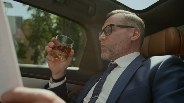 Middle aged Caucasian businessman sitting in backseat of luxury car, drinking whiskey and reading document, low angle shot. Ideal for wealth management and executive travel marketing