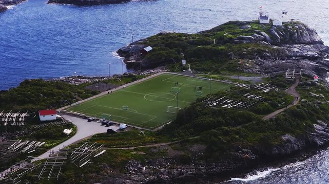 Aerial drone shot shows the famous Henningsvaer football stadium situated on a rocky islet surrounded by the sea with fish drying racks in Lofoten Norway.