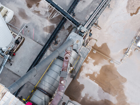 Aerial view of an industrial aggregate processing facility featuring conveyor belts, large metal pipes, and storage silos surrounded by gravel piles in Latvia.