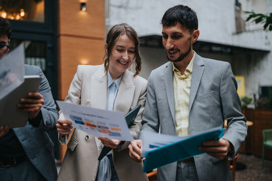 Two business colleagues stand outside reviewing printed documents and smiling as they discuss. The pair examine charts and reports together in a casual outdoor meeting setting.