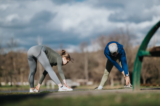 Two girls bend forward stretching their hamstrings and calves on a park path. The girls perform a paired warm-up exercise outdoors before jogging or training.