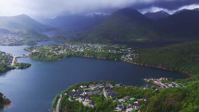 Aerial view over a picturesque coastal town nestled between calm fjord waters and dramatic mountains under cloudy skies in Lofoten, Norway.