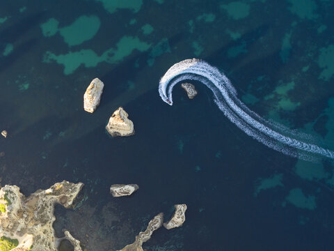 Aerial view of a small boat creating a white wake in the turquoise and deep blue waters of the Atlantic Ocean, surrounded by dramatic limestone rock formations off the coast of Portugal.