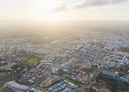 Aerial view of a residential town with scattered buildings, green spaces, and roads under a warm sunset light in Portugal.