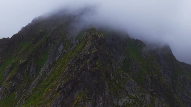 An aerial shot slowly approaches a steep, rocky mountain ridge surrounded by dense fog and clouds in the Lofoten archipelago, Norway.