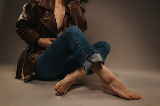 A barefoot woman wearing a brown leather jacket and rolled jeans sits on a studio floor. The casual seated pose emphasizes relaxed style and natural bare feet.