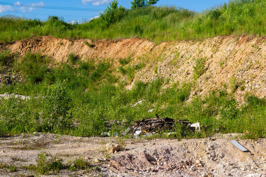 Scattered garbage pile abandoned in an overgrown limestone quarry, featuring assorted debris and waste materials dumped in a raw, natural rocky landscape under a sunny summer sky