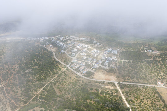 Aerial view of a residential neighborhood with villas and houses nestled among rolling hills and green vegetation, partially covered by low clouds, Portugal.