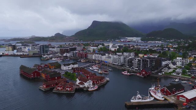 Aerial view of Svolvaer harbour in Lofoten, Norway, showing traditional rorbu cabins, modern buildings, boats, and cloudy mountains.
