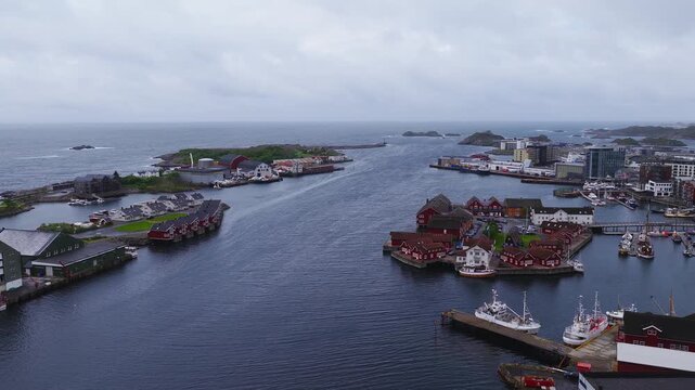 Aerial drone shot over the harbour inlet in Svolvaer, Lofoten, showing waterfront buildings, docks, and moored boats under an overcast sky.