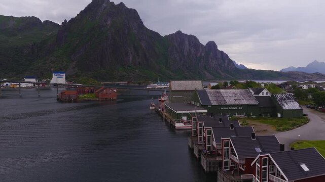 Aerial shot of waterfront cabins, harbour buildings, and steep mountains under an overcast sky in Svolvaer, Lofoten Islands, Norway.