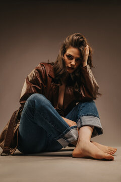 Young woman model sits barefoot on a studio floor wearing a brown leather jacket and rolled jeans. A moody, seated portrait captures a thoughtful, edgy fashion pose in a neutral studio.