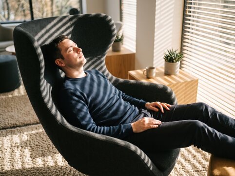 Relaxed man reclining in lounge chair, enjoying warm sunlight through window blinds in modern home interior, creating peaceful atmosphere for rest and comfort