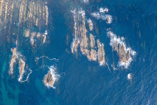 Aerial view of jagged rock formations rising from the deep blue ocean with white sea foam crashing against the rugged stone surfaces in Portugal.