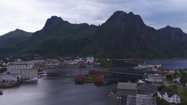 Aerial view shows the coastal town of Svolvaer in Lofoten Norway, with boats in the harbour, a road bridge, and steep mountains under cloudy skies.