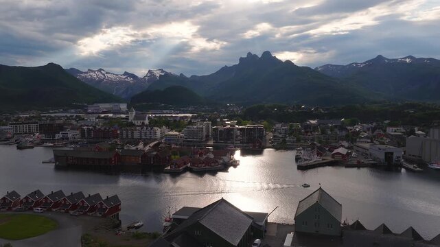 Aerial view over the coastal town and harbour of Svolvaer in Lofoten, Norway, with waterfront cabins, boats, and mountain peaks under cloudy sky.